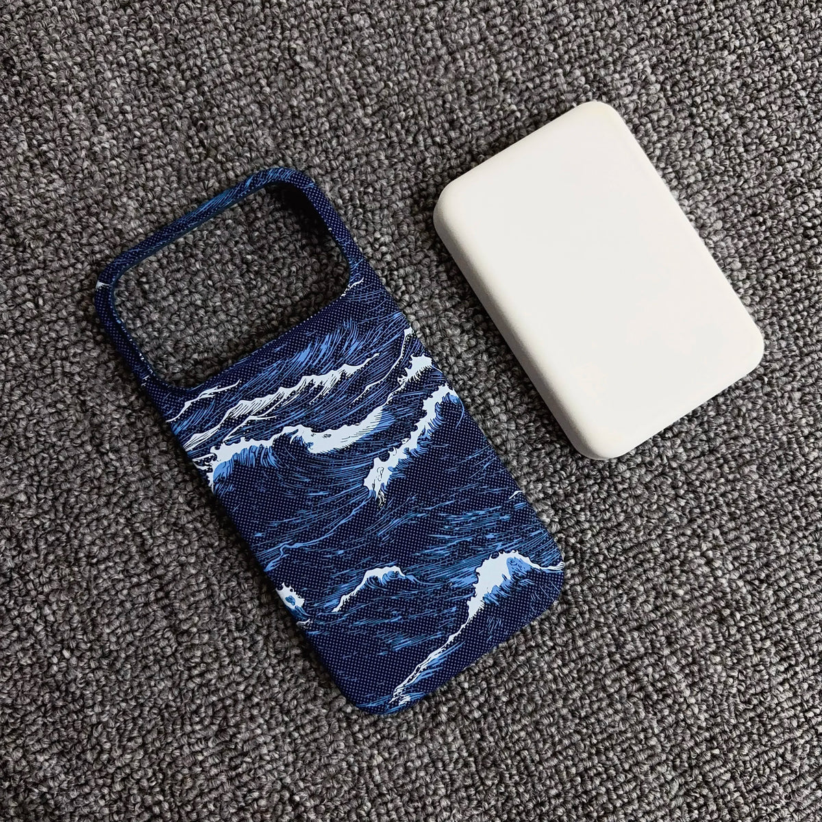 A phone case with a blue and white ocean wave pattern, shown next to a white rectangular power bank on a textured gray carpet.