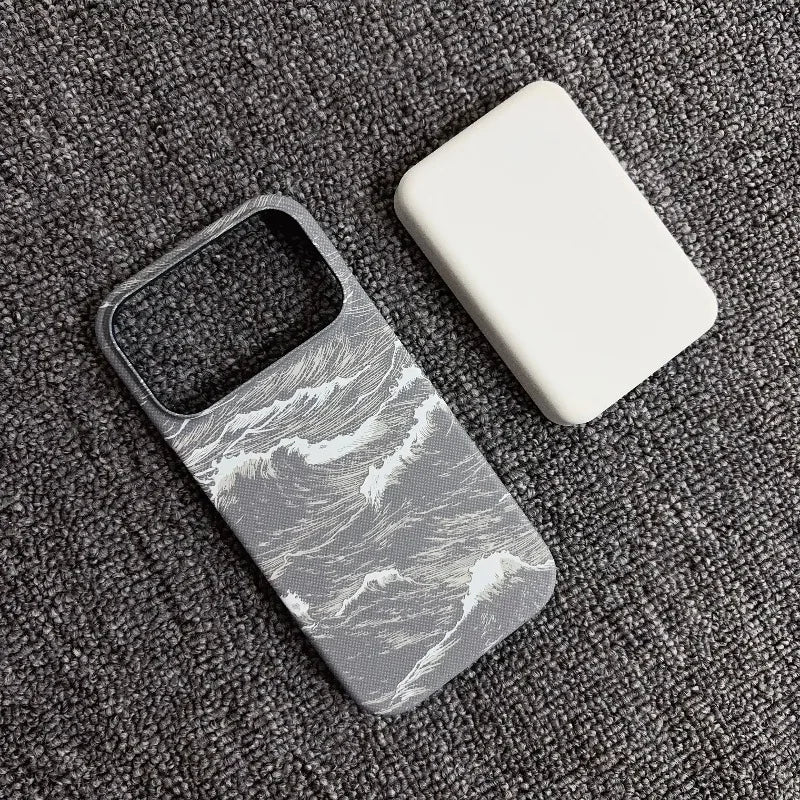 A gray and white ocean wave pattern phone case next to a plain white rectangular protective cover, displayed on a dark gray textured fabric background.