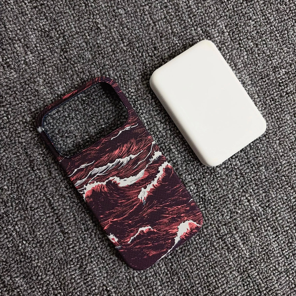 A phone case with a dark red and black marble wave pattern, shown next to a white rectangular protective cover on a textured gray carpet.