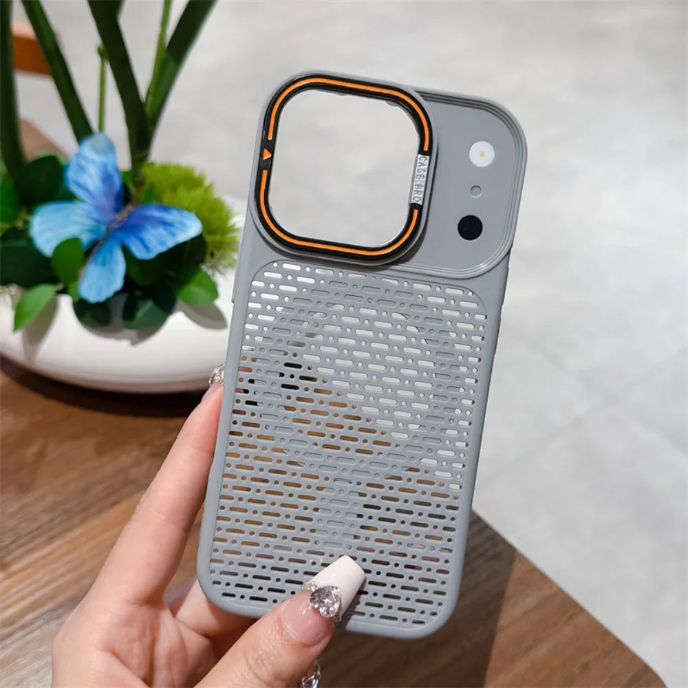 A hand holds a gray perforated phone case with a raised camera bezel and orange accent trim, photographed indoors with a blurred plant and tabletop in the background.