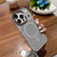A hand holding a gray textured phone case with a circular speaker-like grille and a raised camera surround outlined in orange, photographed over a wooden table with a blurred drink and plant in the background.