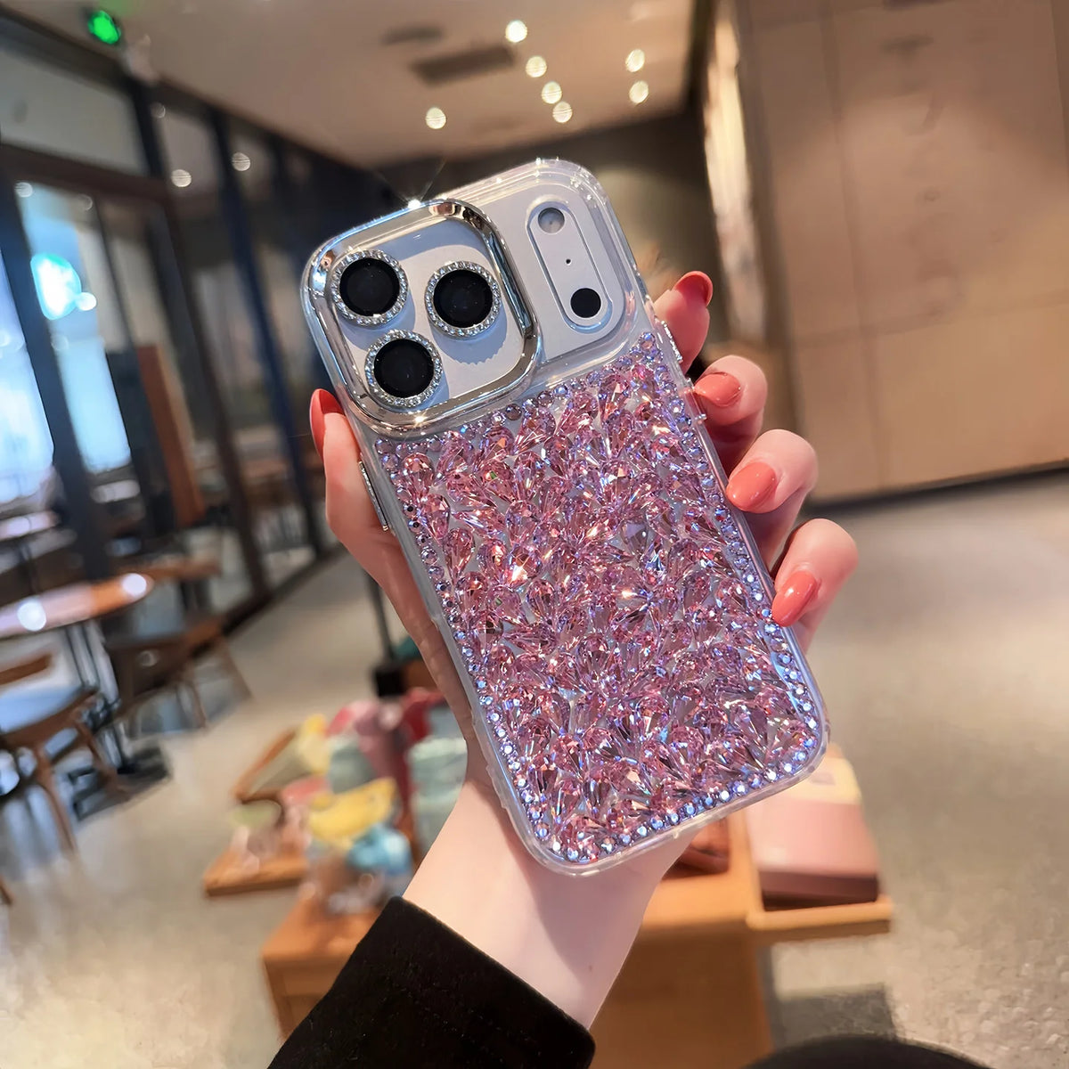 A hand holding a clear phone case covered in pink and silver rhinestones, with a gold-toned camera rim, shown in a blurred indoor setting.