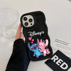 A hand holding a black phone case featuring Disney's Stitch and Angel with pink hearts, shown on a white tabletop with a clear cup and printed cards nearby.
