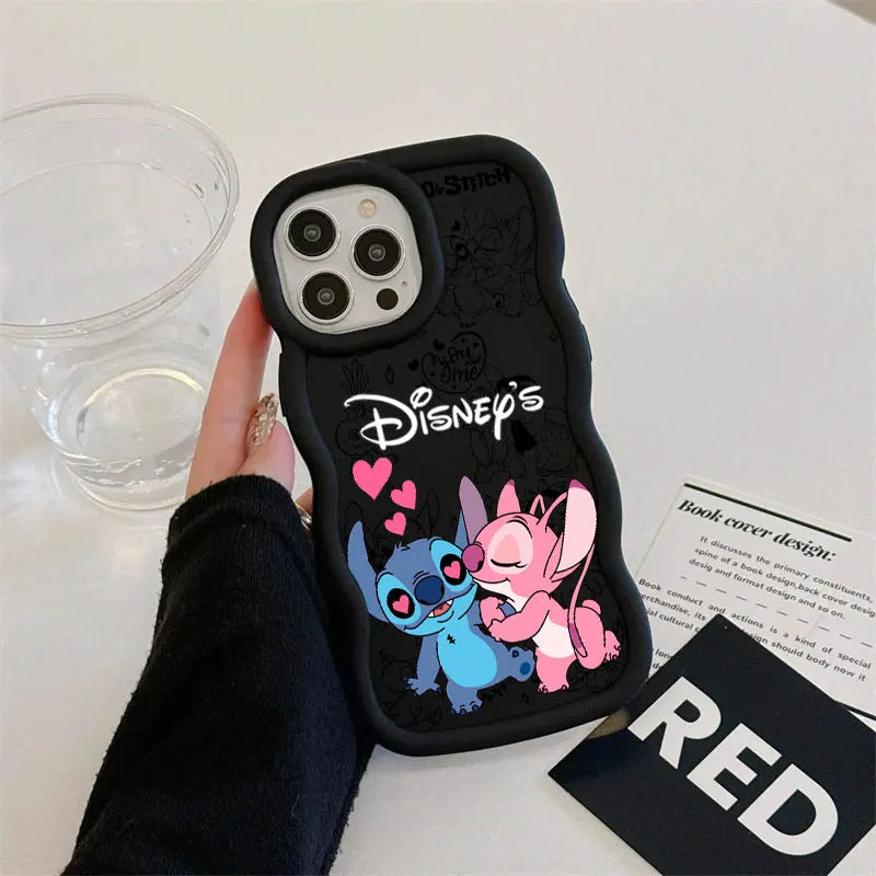 A hand holding a black phone case featuring Disney's Stitch and Angel with pink hearts, shown on a white tabletop with a clear cup and printed cards nearby.
