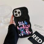 A hand wearing a black sleeve holds a black phone case featuring cartoon characters Stitch and Angel with the word 'Stitch' in stylized lettering. The case is photographed on a white tabletop with a clear plastic cup, a printed card, and a black card with the word 'RED' nearby.