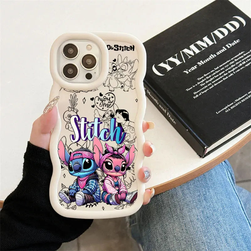 A hand holds a white phone case featuring Stitch and Angel illustrations with the word 'Stitch' in blue. The case is shown in a lifestyle setting next to a black book on a light-colored table.