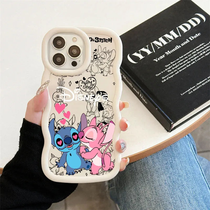 A hand holds a white phone case featuring cartoon characters Stitch and Angel with pink hearts and black doodle graphics. The case is shown on a phone, with a black book and blue jeans in the background.